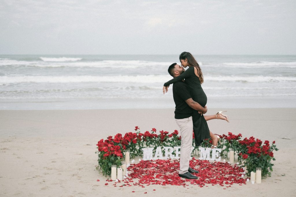 Hombre le propone matrimonio a mujer en la playa con rosas y velas.